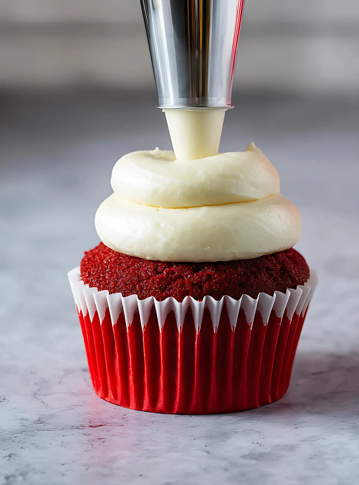 Piping bag adding white frosting to red velvet cupcake